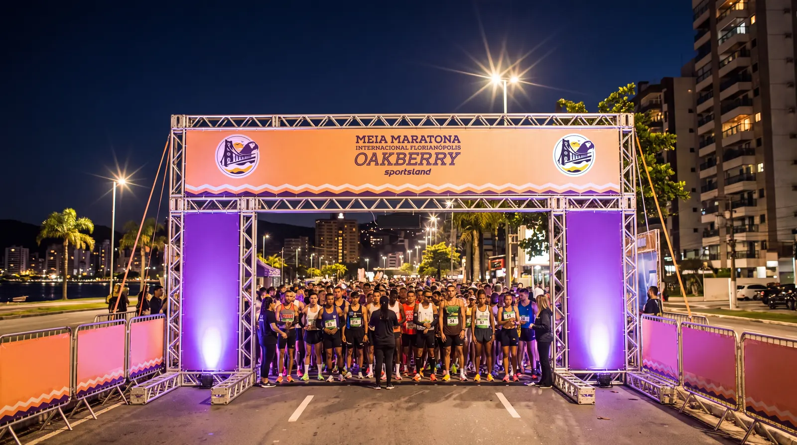 Pórtico de la carrera encendido de madrugada, columnas violetas y cabecera salmón, con corredores calentando en el corral